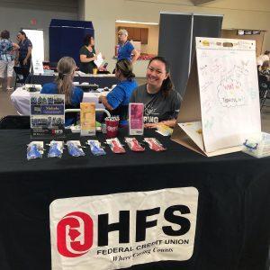 hfs staff member smiles from behind a table at the hmsa akamai living fair. the table, covered in a black tablecloth with the HFS logo in front, has various brochures and promotional items on top